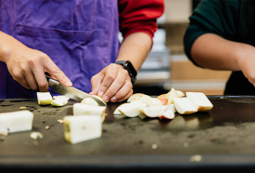 You can see the hands of a student, working in a classroom kitchen and cutting into an apple. Another student is partially visible in the background.